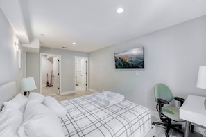 Ein modernes Hotelzimmer mit einem großen Bett, einem Schreibtisch und einem Sessel. An der Wand hängt ein Gemälde, und eine Tür führt in einen weiteren Raum.