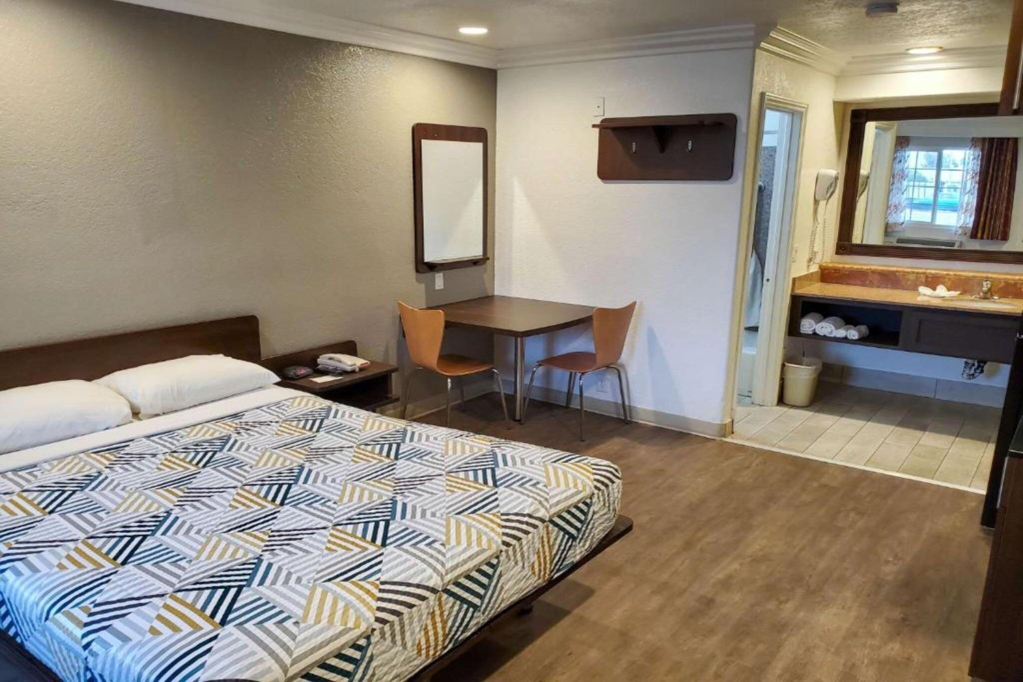 Habitación de hotel con cama, escritorio, silla, espejo y baño con lavamanos e inodoro.