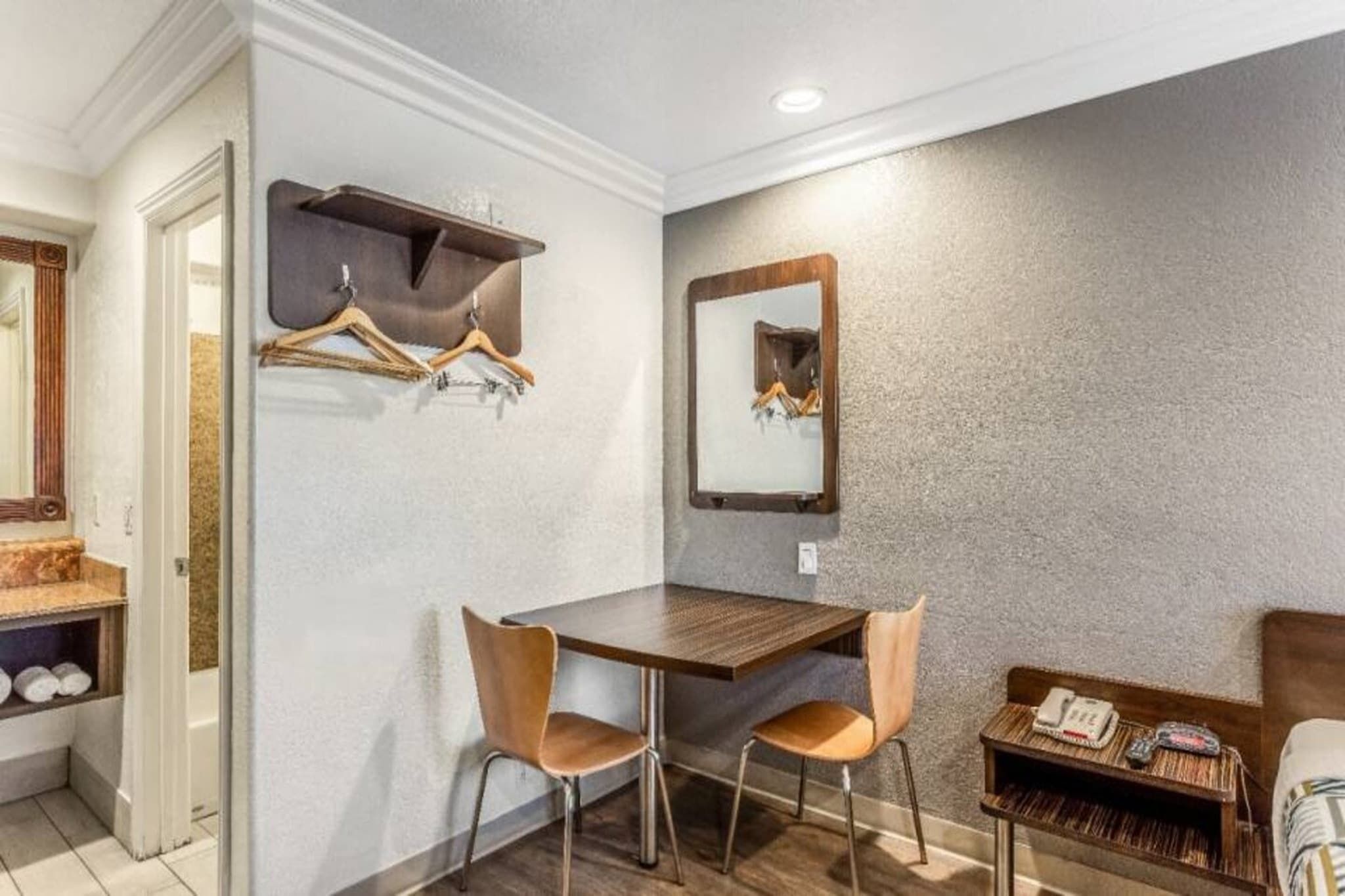 Ein Hotelzimmer mit einem Holztisch, zwei Stühlen, einem Spiegel und einem Kleiderhaken.