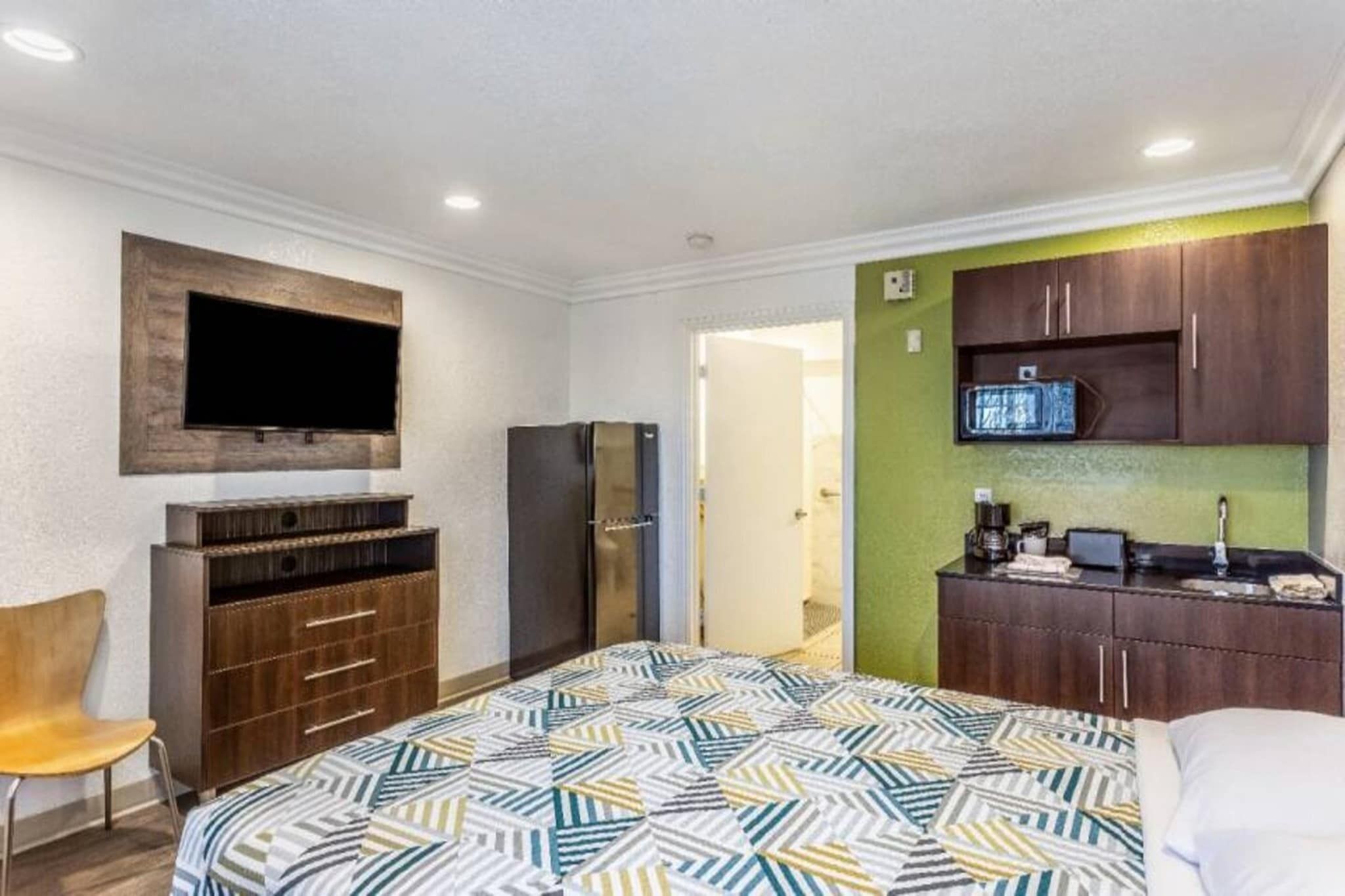 A hotel room with a bed, a TV mounted on the wall, a refrigerator, a wooden cabinet, and a kitchenette area.