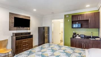A hotel room with a bed, a TV mounted on the wall, a refrigerator, a wooden cabinet, and a kitchenette area.