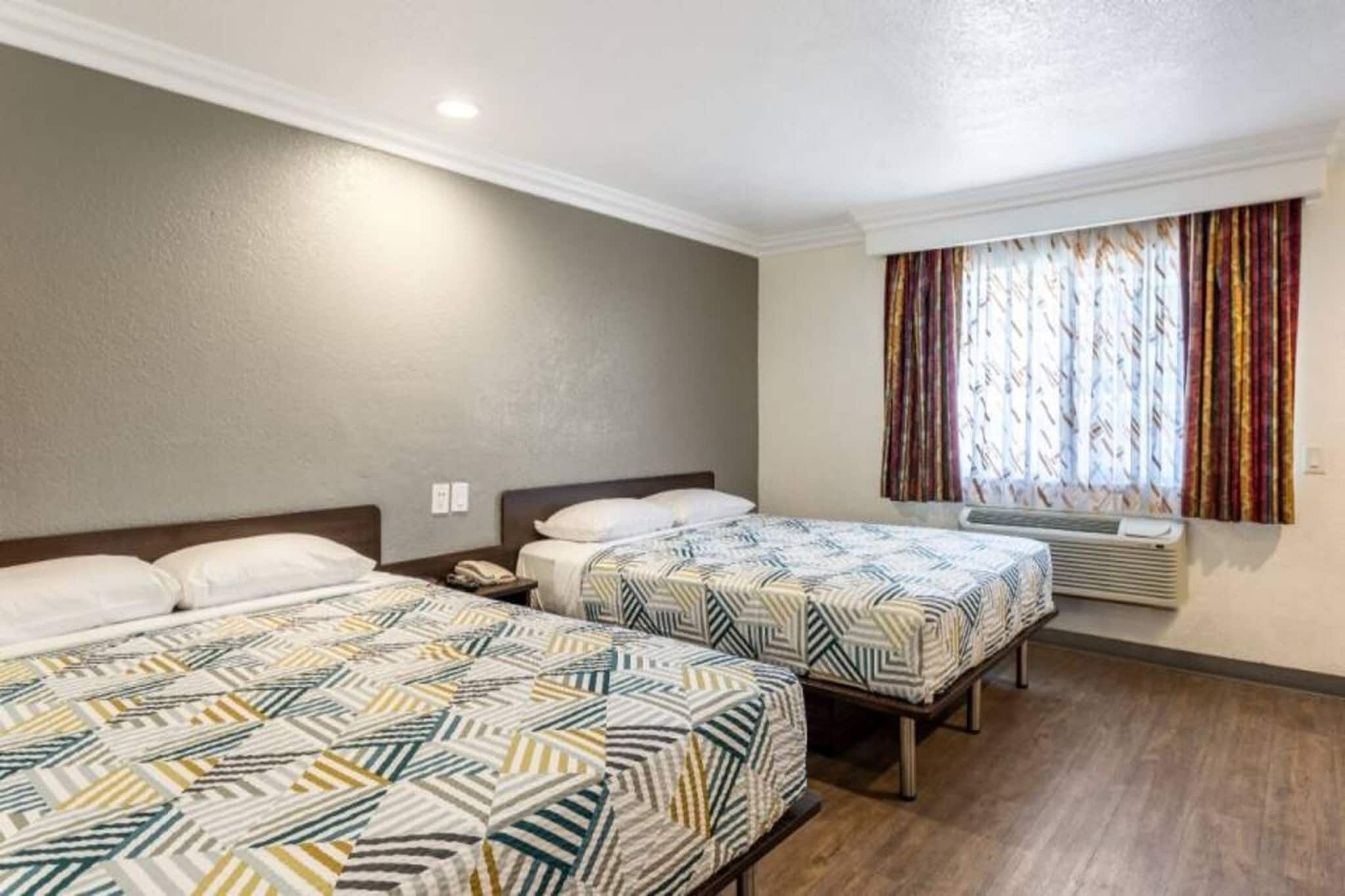 A hotel room with two beds, a window with curtains, and a wall-mounted air conditioner.