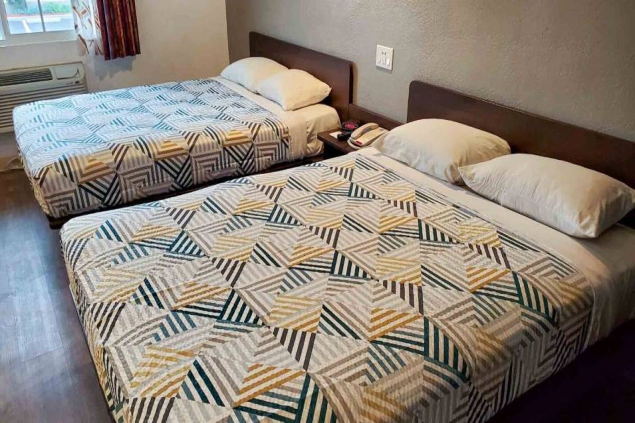 Two double beds with patterned bedding in a room with a window and a wall socket.