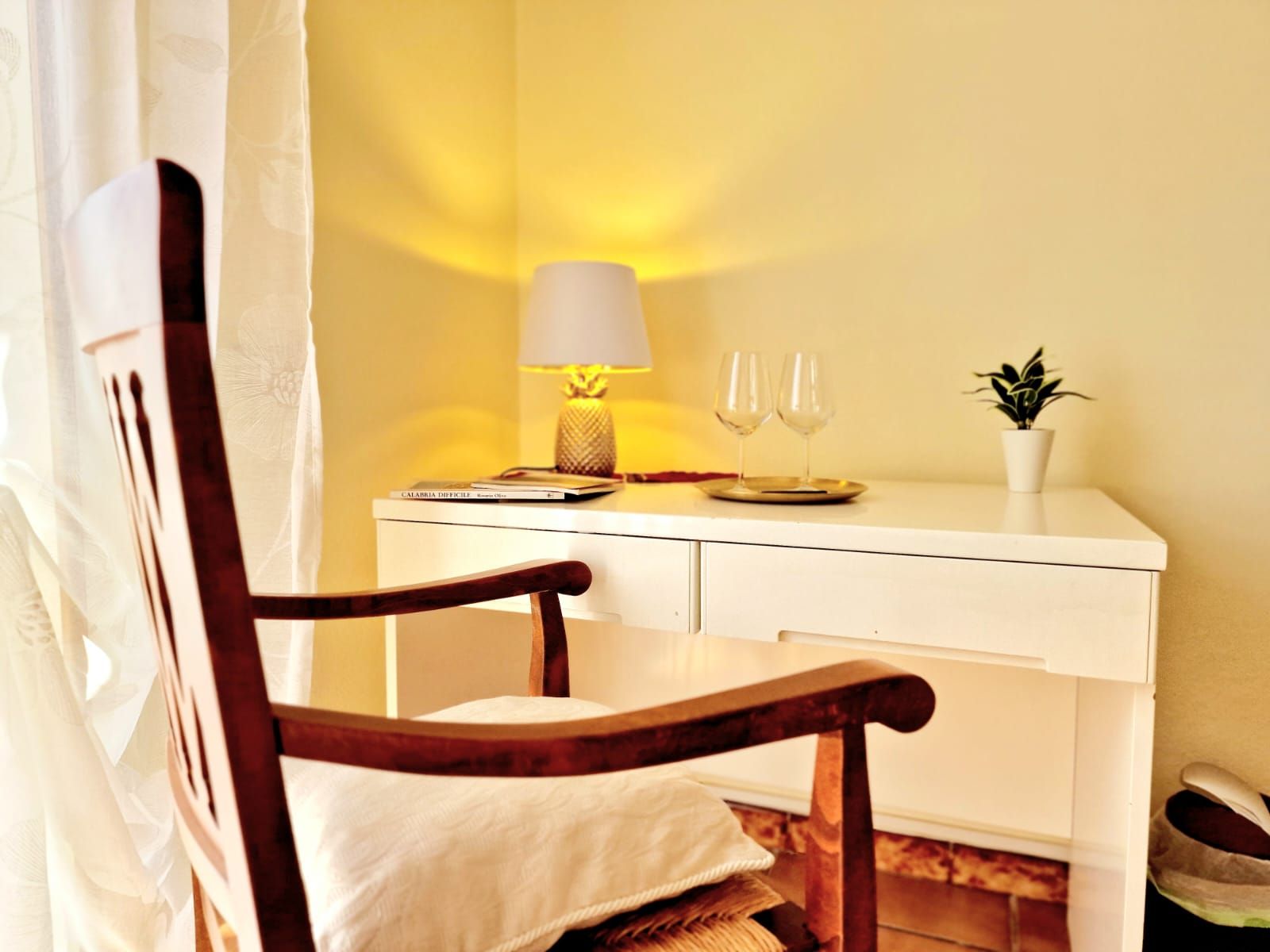 Ein Zimmer mit einem Holzstuhl, einem weißen Schreibtisch, einer Lampe, zwei Weingläsern und einer kleinen Topfpflanze.