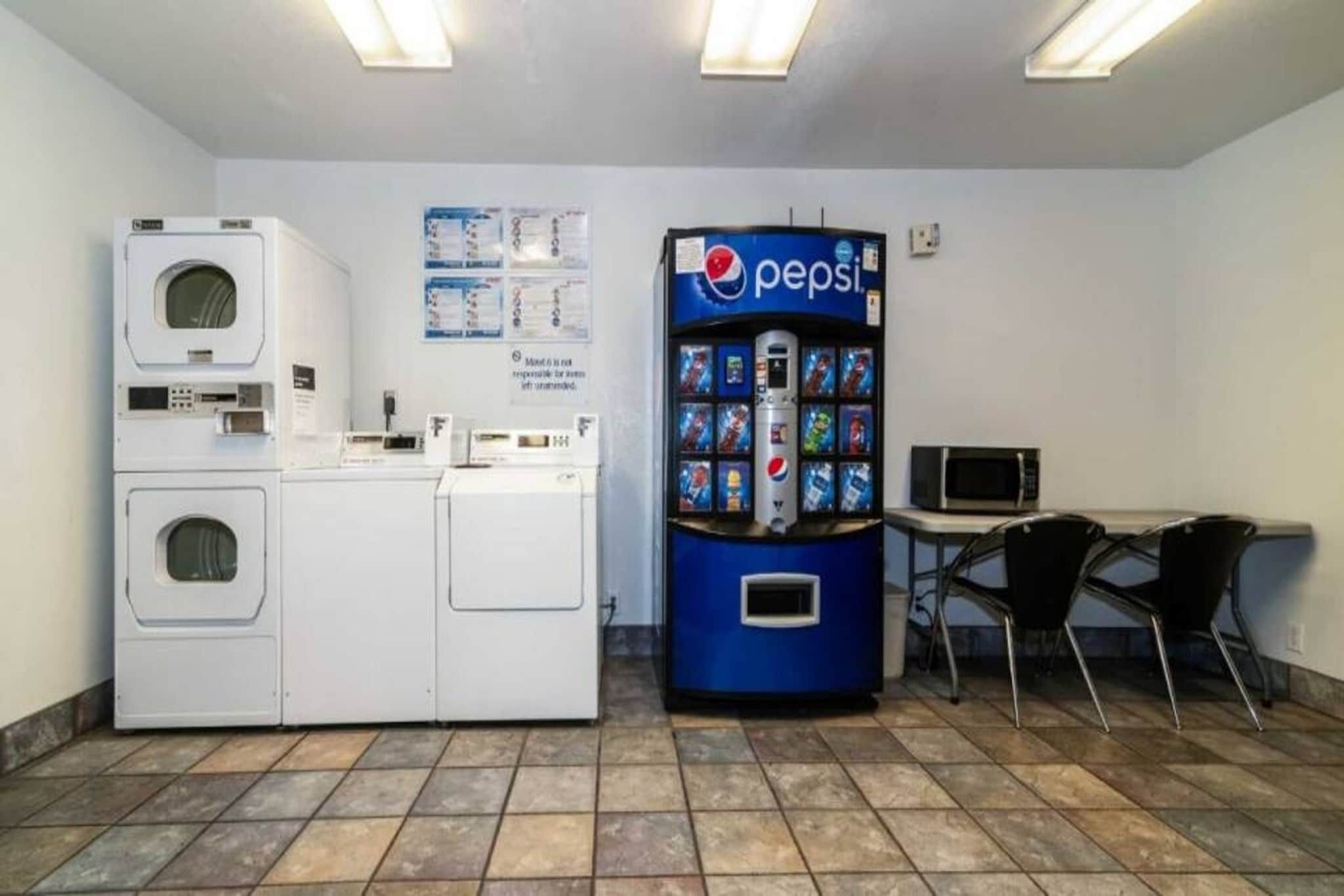 Uma área de lavanderia com máquinas de lavar e secar, um micro-ondas e uma máquina de refrigerantes Pepsi.