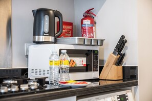 Uma bancada de cozinha com chaleira, extintor de incêndio, micro-ondas, conjunto de facas e um prato com comida.