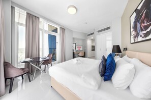 A hotel room with a bed, a desk, a chair, a window with curtains, and a painting on the wall. - StayShort - Cozy Studio in Damac Hills 1 for 2 with Pool Access! (Damac HIlls 1)