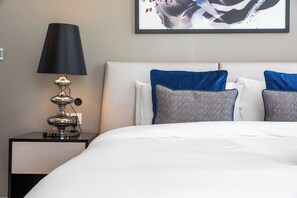 Ein ordentlich bezogenes Bett mit weißer Bettwäsche, eine schwarze Lampe auf einem Nachttisch und zwei Kissen mit blauem Muster.