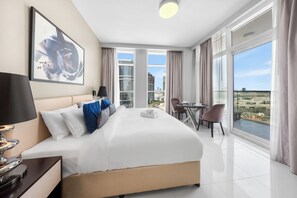 A hotel room with a large bed, a desk, a chair, and a view of the cityscape. - StayShort - Cozy Studio in Damac Hills 1 for 2 with Pool Access! (Damac HIlls 1)