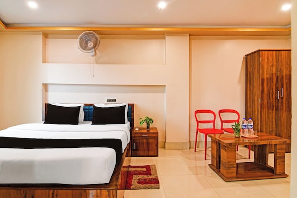A hotel room with a bed, bedside table, nightstand, a fan, a wooden wardrobe, a small table with chairs, and a water bottle. - Collection O Kalyani Expressway Kolkata (Barakpur)