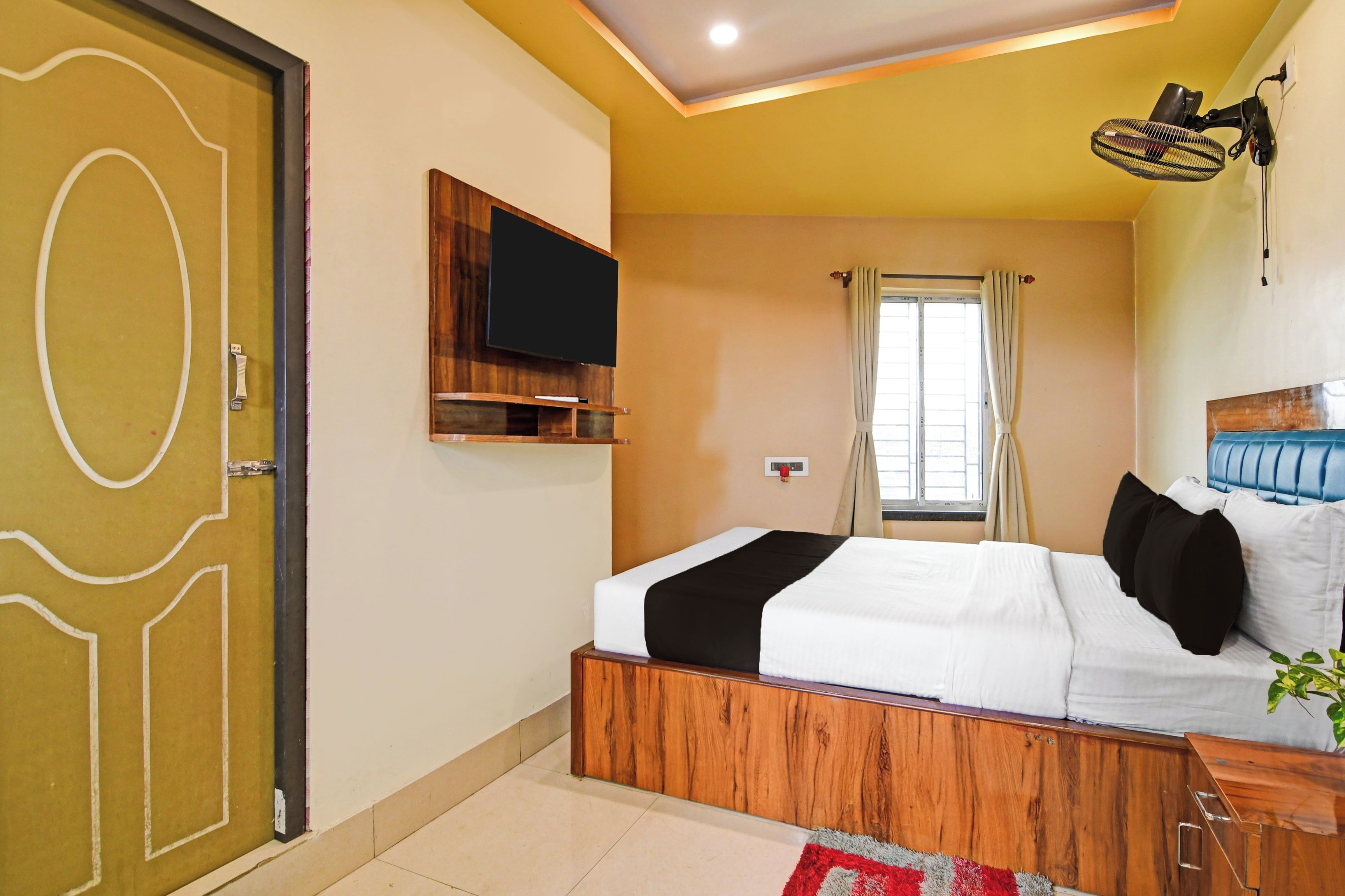 A hotel room with a bed, a TV mounted on the wall, a window with curtains, and a ceiling fan.