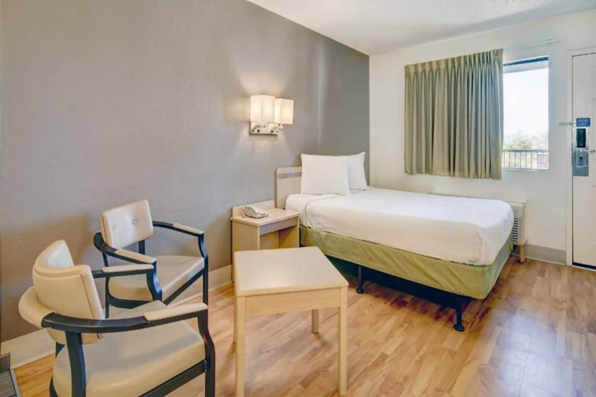 A hotel room with a bed, a chair, a small table, a nightstand, a lamp, and a window with curtains.