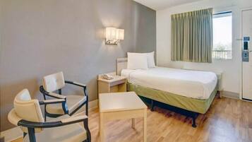 A hotel room with a bed, a chair, a small table, a nightstand, a lamp, and a window with curtains.