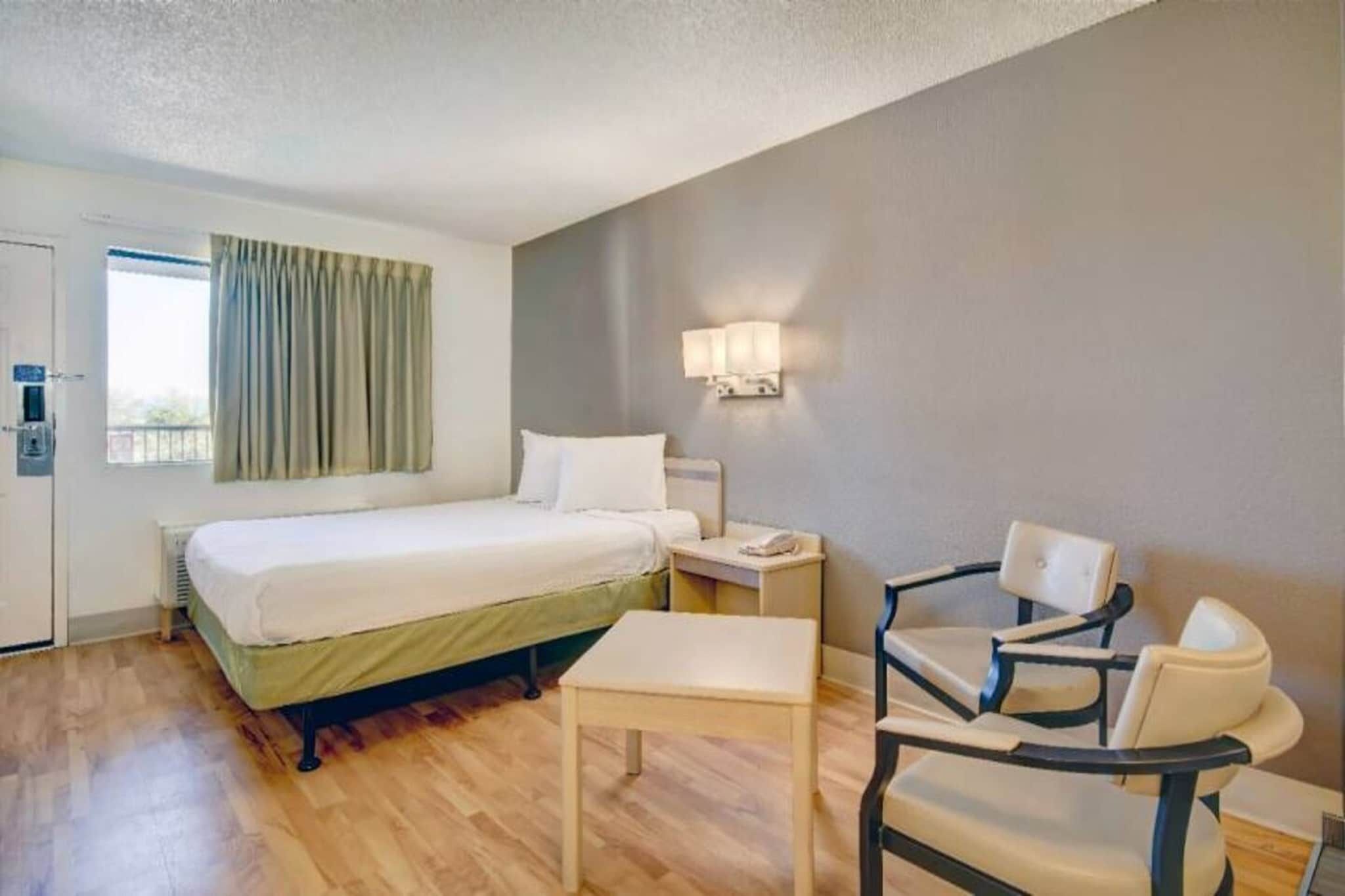 Quarto de hotel com cama, mesa de cabeceira, cadeira e janela com cortinas.