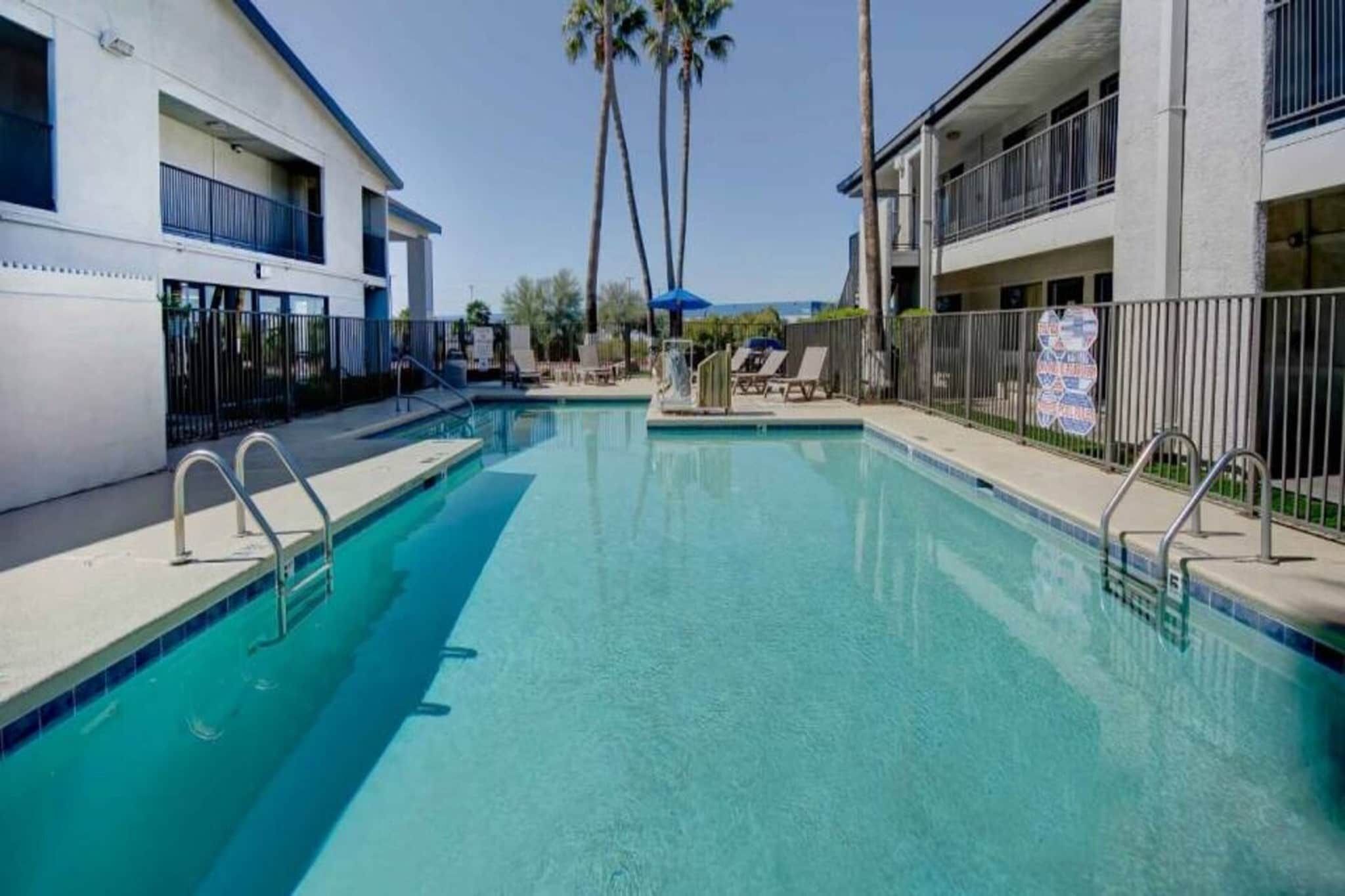 A swimming pool with a clear blue water, surrounded by a white building with balconies and palm trees in the background.