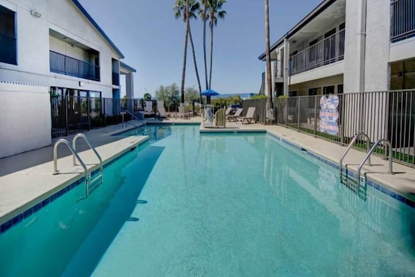 Una piscina con agua azul cristalina, rodeada por un edificio blanco con balcones y palmeras al fondo.