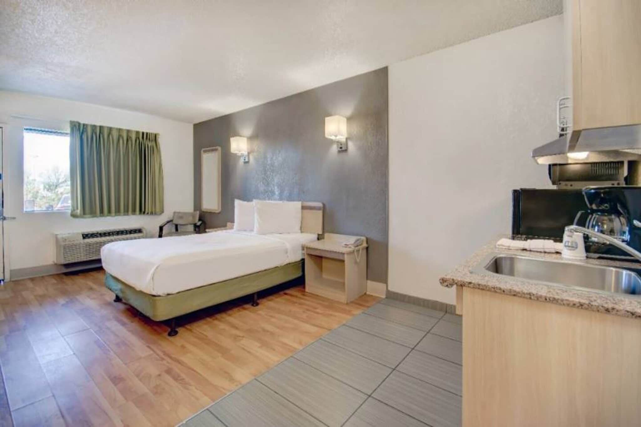 Une chambre d’hôtel comprenant un lit, une table de chevet, un petit bureau, une chaise, une fenêtre avec des rideaux, un miroir fixé au mur, un radiateur et un coin cuisine avec un évier et une machine à café.