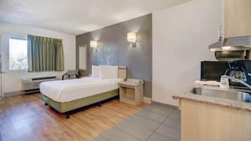 Ein Hotelzimmer mit Bett, Nachttisch, kleinem Schreibtisch, Stuhl, Fenster mit Vorhängen, wandmontiertem Spiegel, Heizung und einer Küchenzeile mit Spüle und Kaffeemaschine.