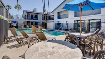 A pool area with lounge chairs and umbrellas, a round table, and a building with balconies and palm trees in the background.