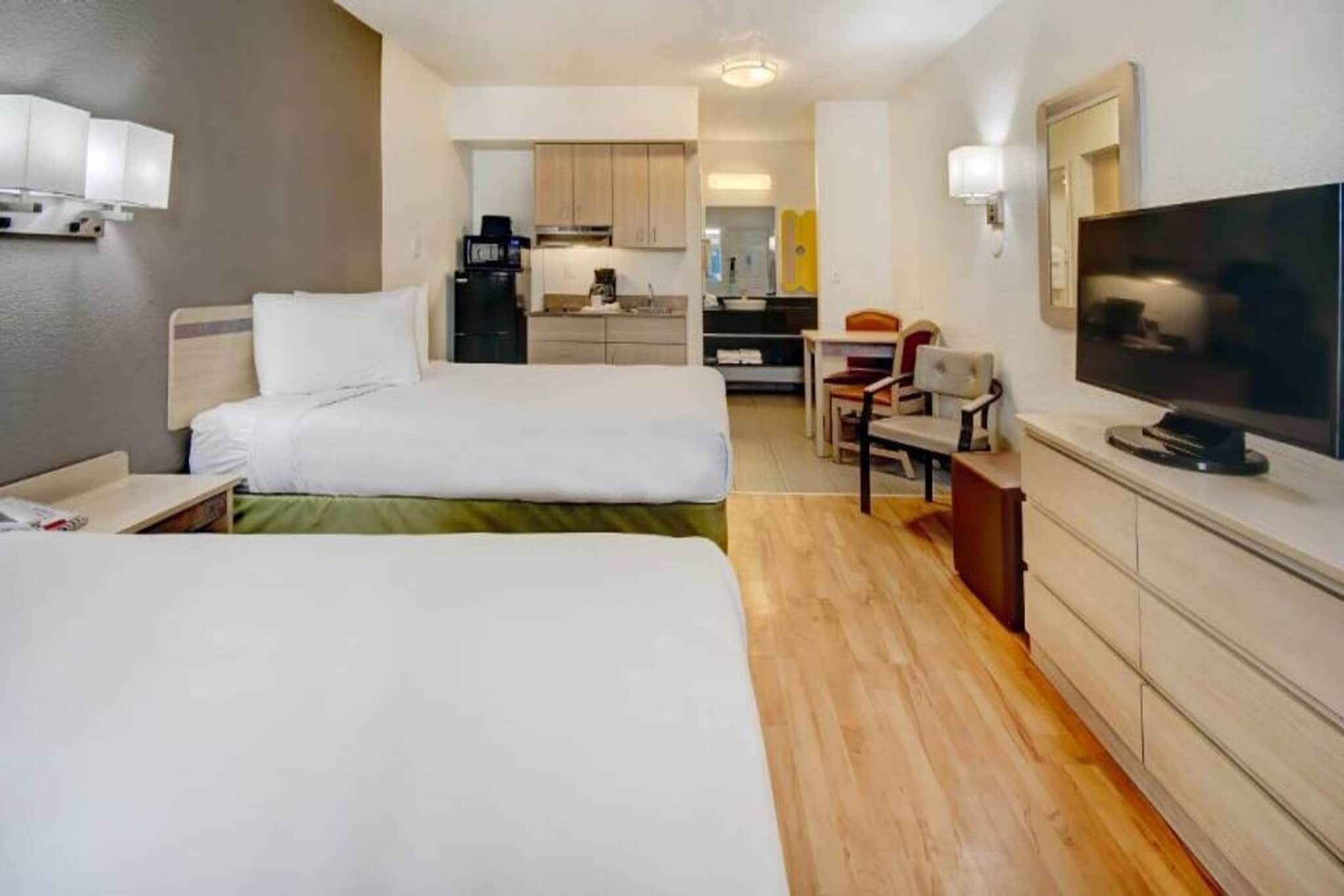A hotel room with two beds, a television, a desk, and a kitchenette in the background.