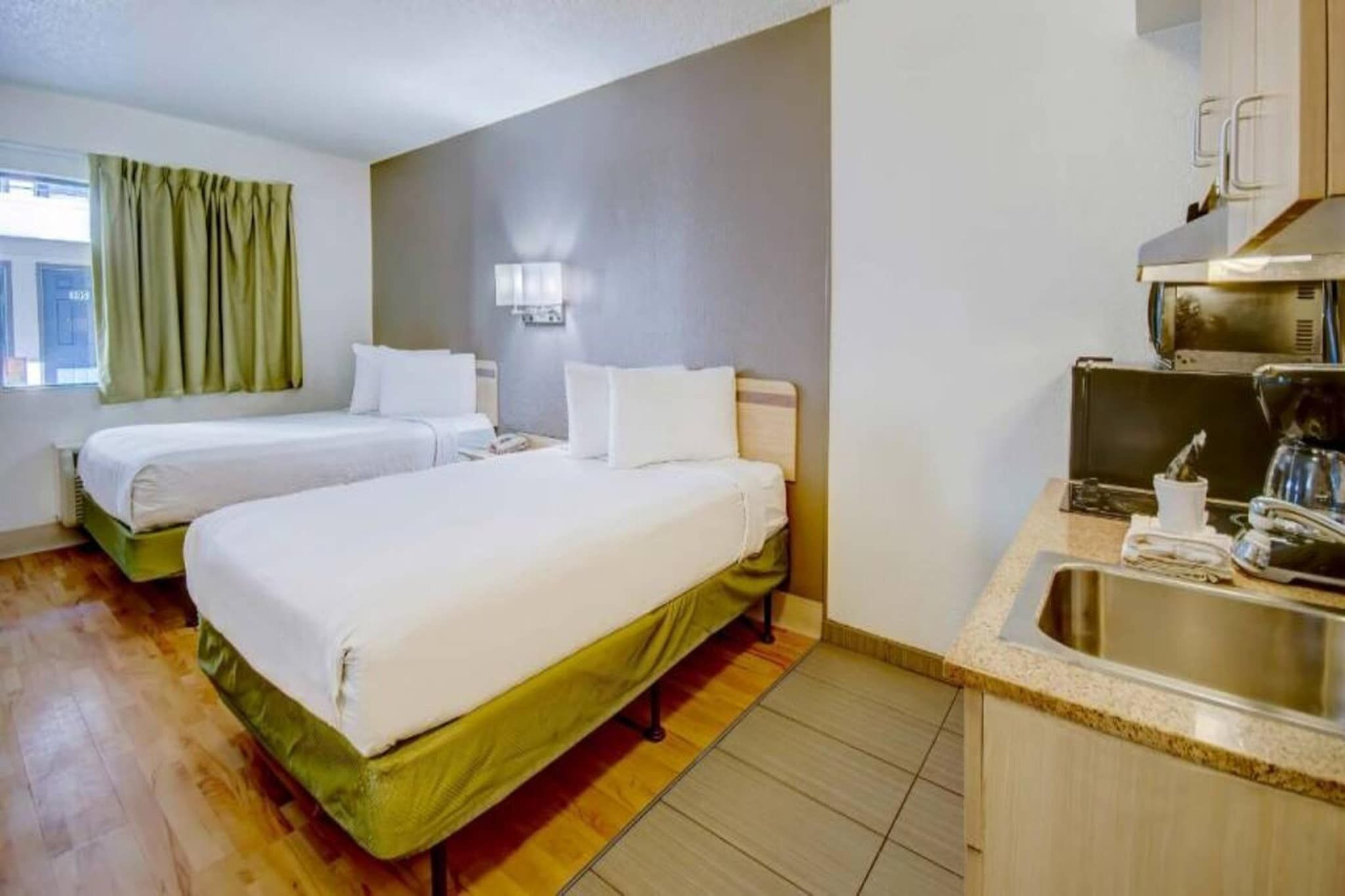 Ett hotellrum med två sängar, ett litet kök och ett fönster med gardiner.