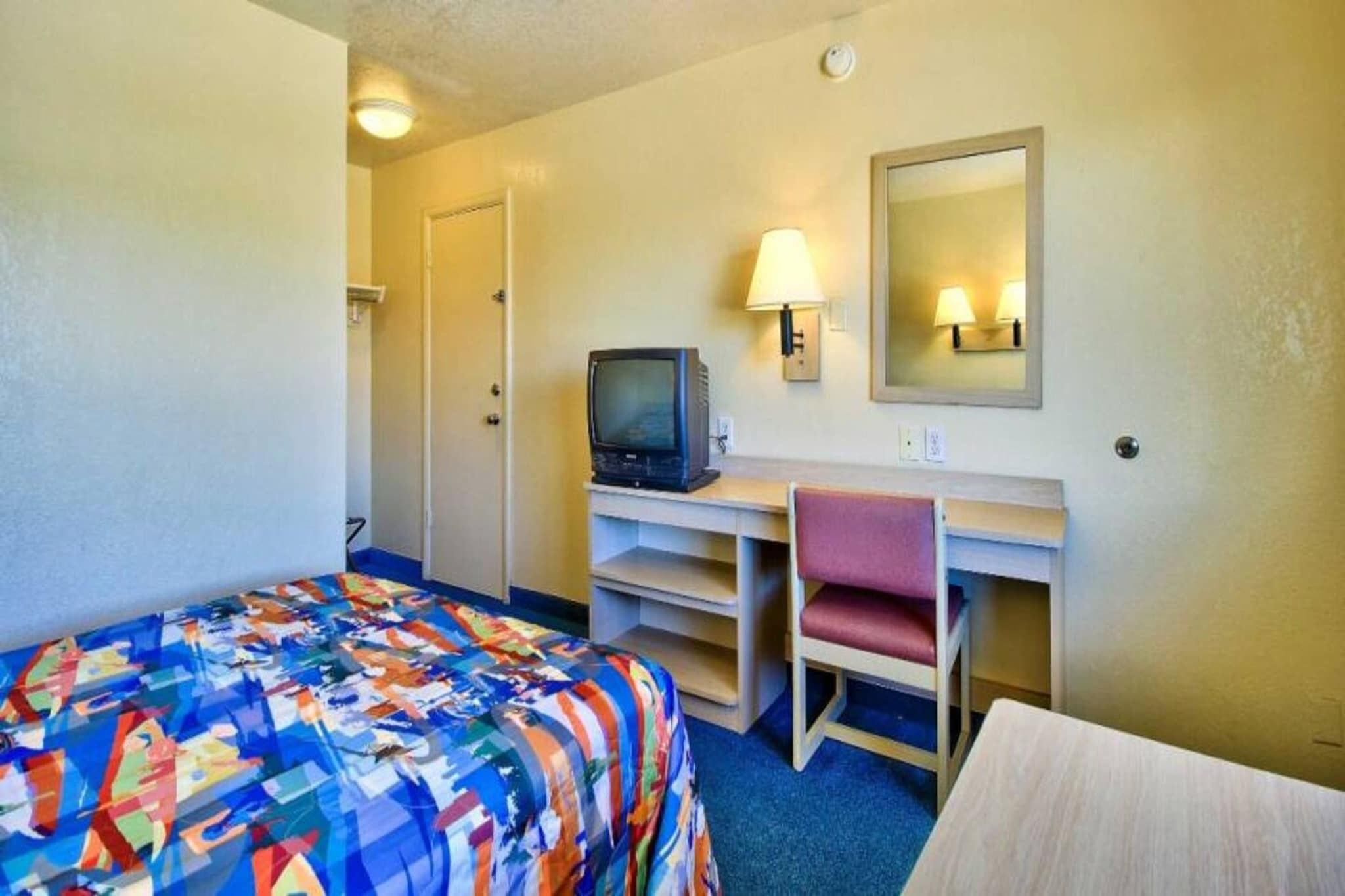 Una habitación de hotel con cama, escritorio, silla, televisión y espejo.