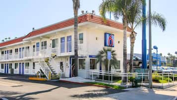 A two-story motel with a red-tiled roof, multiple rooms with blue doors, and a parking area with a Chevrolet dealership in the background.