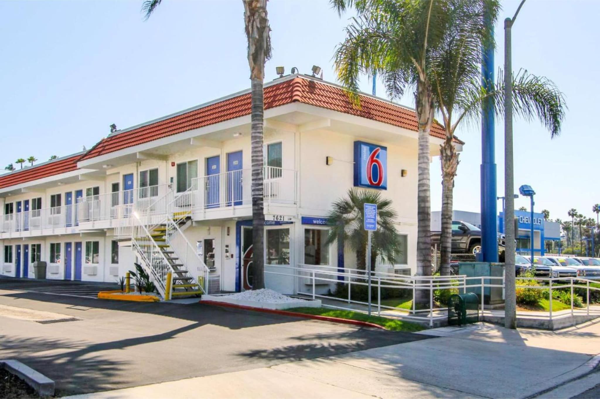 Un motel à deux étages, avec un toit recouvert de tuiles rouges, de nombreuses chambres dotées de portes bleues et un parking où l’on aperçoit, au fond, une concessionnaire Chevrolet.