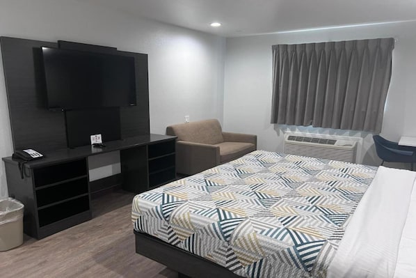 Habitación de hotel con cama, televisión montada en la pared, una silla, escritorio y ventana con cortinas.