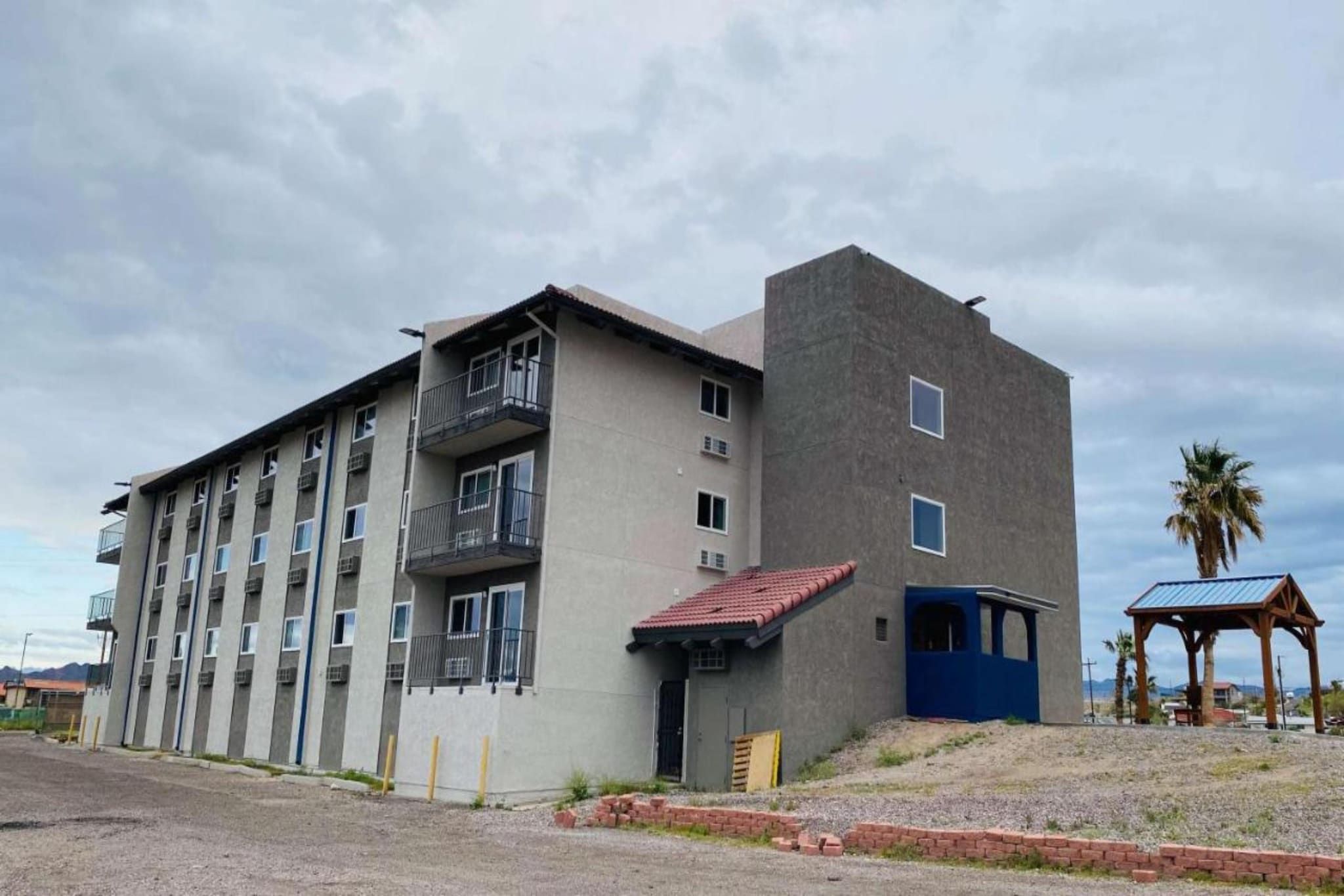 Un edificio de varios pisos con balcones, una estructura pequeña adosada con una puerta azul, y un área cubierta con techo.