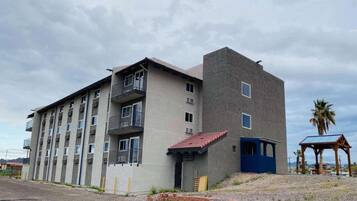 Un edificio de varios pisos con balcones, una estructura pequeña adosada con una puerta azul, y un área cubierta con techo.