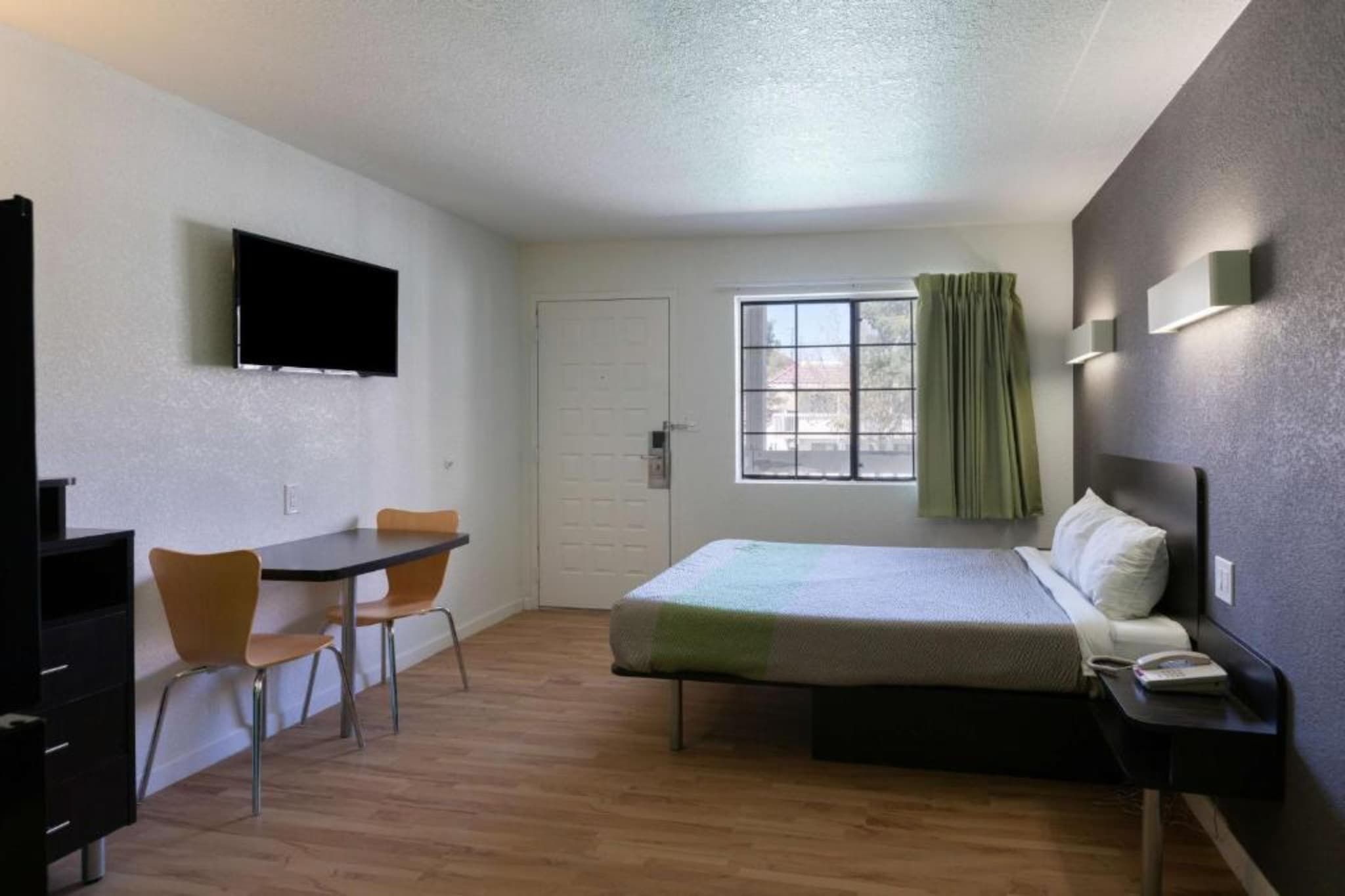 A small, clean room with a bed, a desk, a chair, a television, and a window with curtains.