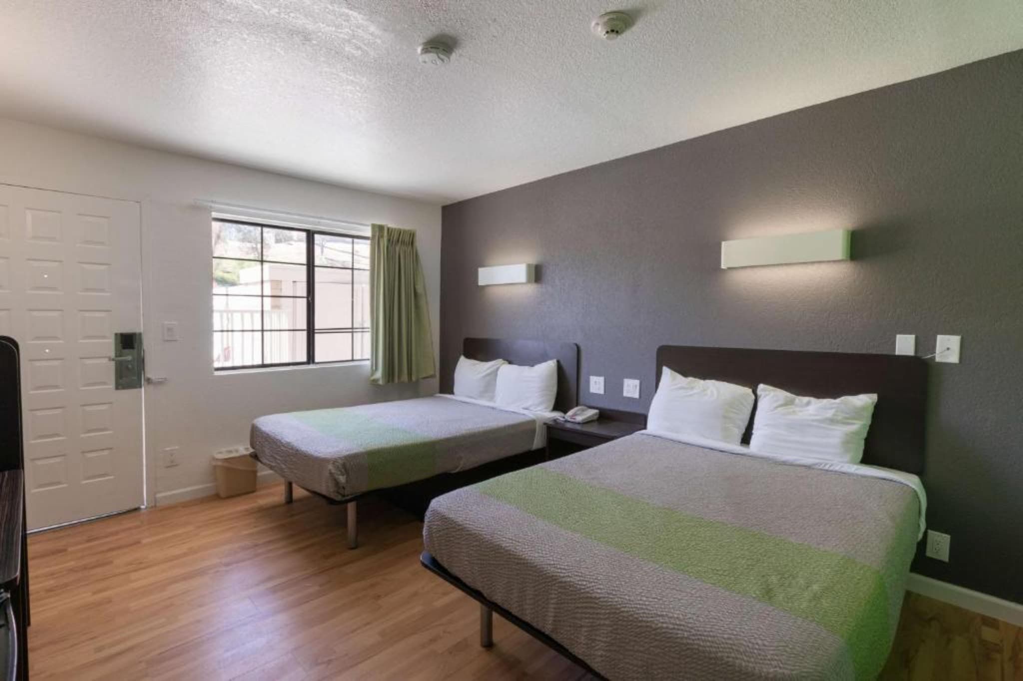 A hotel room with two single beds, a window with curtains, a door, and a light switch on the wall.