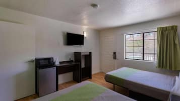 A small, simple room with a desk, a television mounted on the wall, a bed, and a window with curtains.