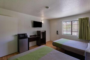 Habitació petita i senzilla amb escriptori, televisió penjada a la paret, llit i finestra amb cortines.