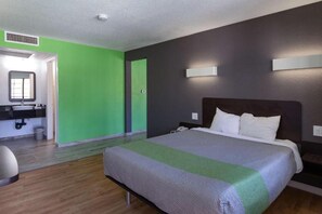 Habitación de hotel con cama, mesita de noche, espejo y baño con lavamanos.