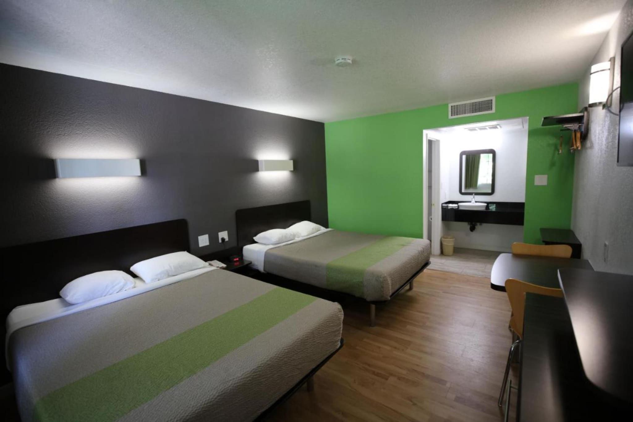 Habitación de hotel con dos camas, un baño con espejo y lavamanos, y una mesita pequeña.