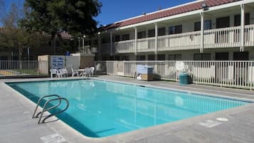 A swimming pool with a metal ladder, surrounded by a white fence, and a building with balconies in the background.
