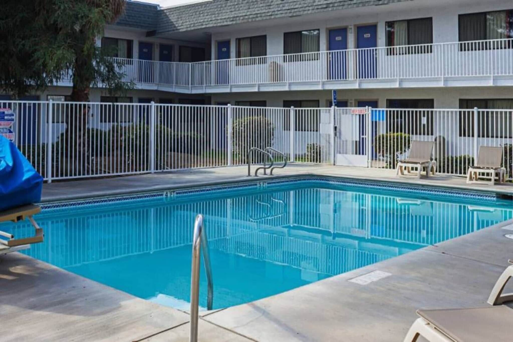 A swimming pool with a fenced deck, lounge chairs, and a multi-story building in the background.