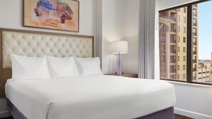 Een hotelkamer met een groot bed, een hoofdbord, een nachtkastje met een lamp, een raam met uitzicht op gebouwen en een schilderij aan de muur.