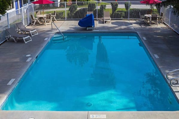 A rectangular swimming pool with a 5 ft depth marker, surrounded by a white fence, and equipped with lounge chairs and umbrellas.