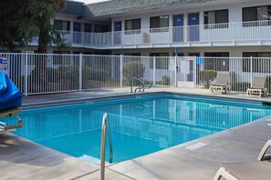 A swimming pool with a fenced deck, lounge chairs, and a multi-story building in the background.
