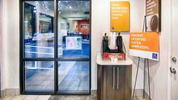 A hotel reception area with a glass door entrance, a coffee station, and informational signs about room rates and updates.