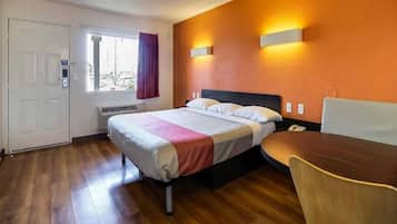 A hotel room with a bed, a desk, and a chair. There is a window with curtains, a door, and a wall-mounted light fixture.