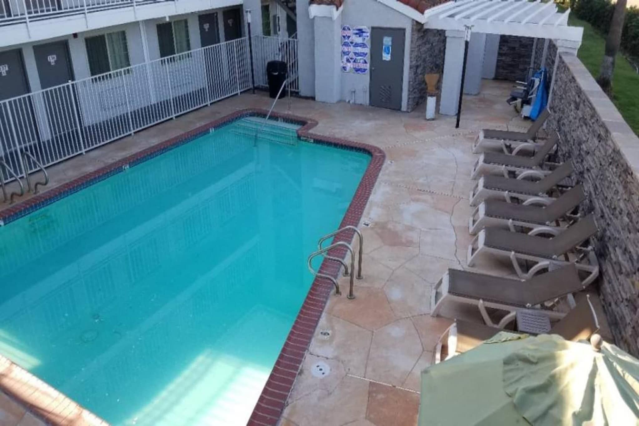 A swimming pool with a tiled deck, a covered area with lounge chairs, and a building with a white railing in the background.