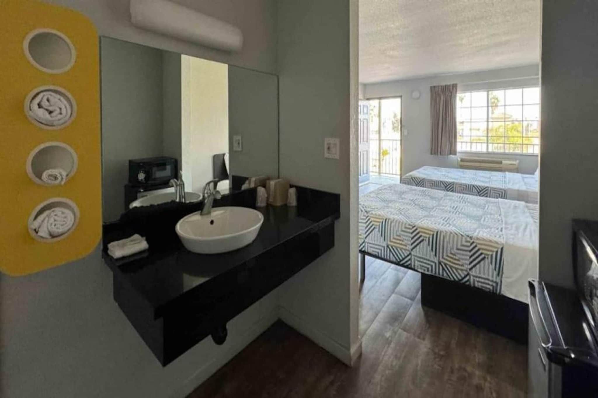 Ein Hotelzimmer mit schwarzer Arbeitsplatte, Waschbecken in Weiß, einem Spiegel und einem Bett mit gemusterten Textilien.