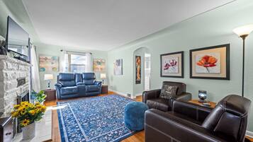A living room with a fireplace, blue and brown leather chairs, a blue patterned rug, and artwork on the walls.