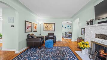 A living room with a fireplace, a blue patterned rug, and a dining area in the background.