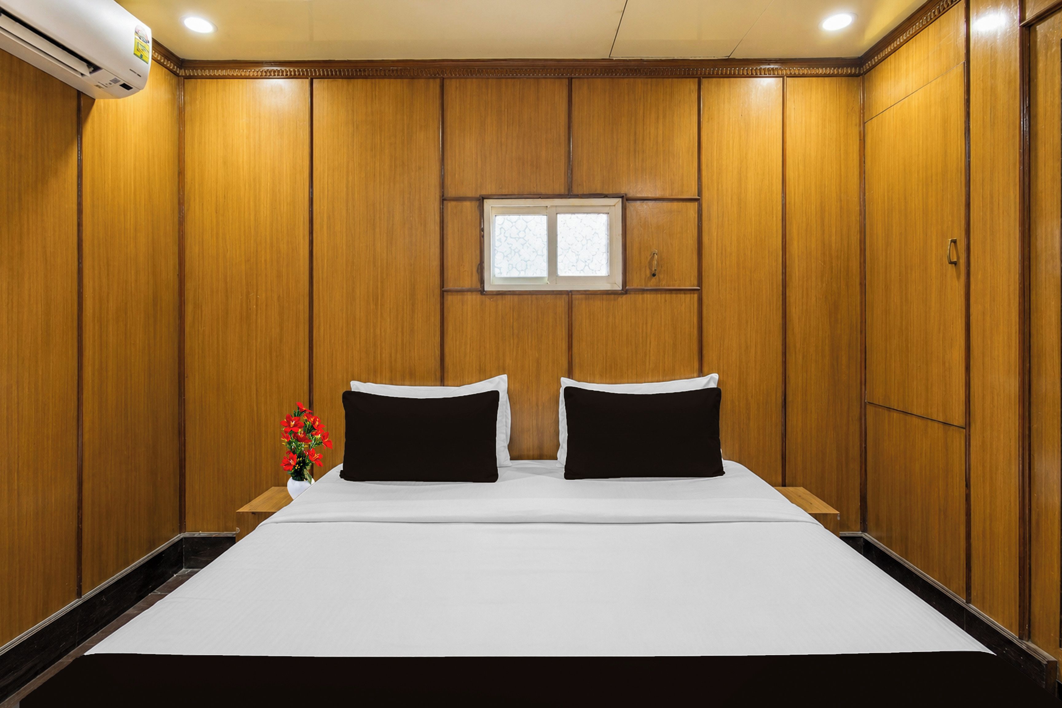 A hotel room with wooden panel walls, a bed with white bedding, two black pillows, a small window, and a wall-mounted air conditioning unit.