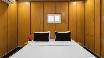Habitación de hotel con paneles de madera, una cama con ropa blanca, dos almohadas negras, un ventanal pequeño y un equipo de aire acondicionado instalado en la pared.